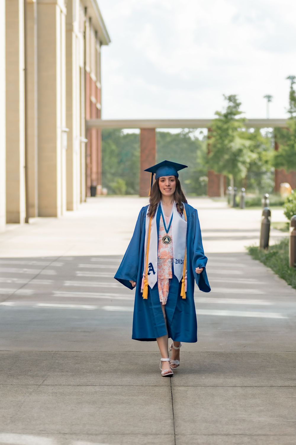 Graduate in blue cap and gown walks confidently along campus sidewalk during commencement ceremony.