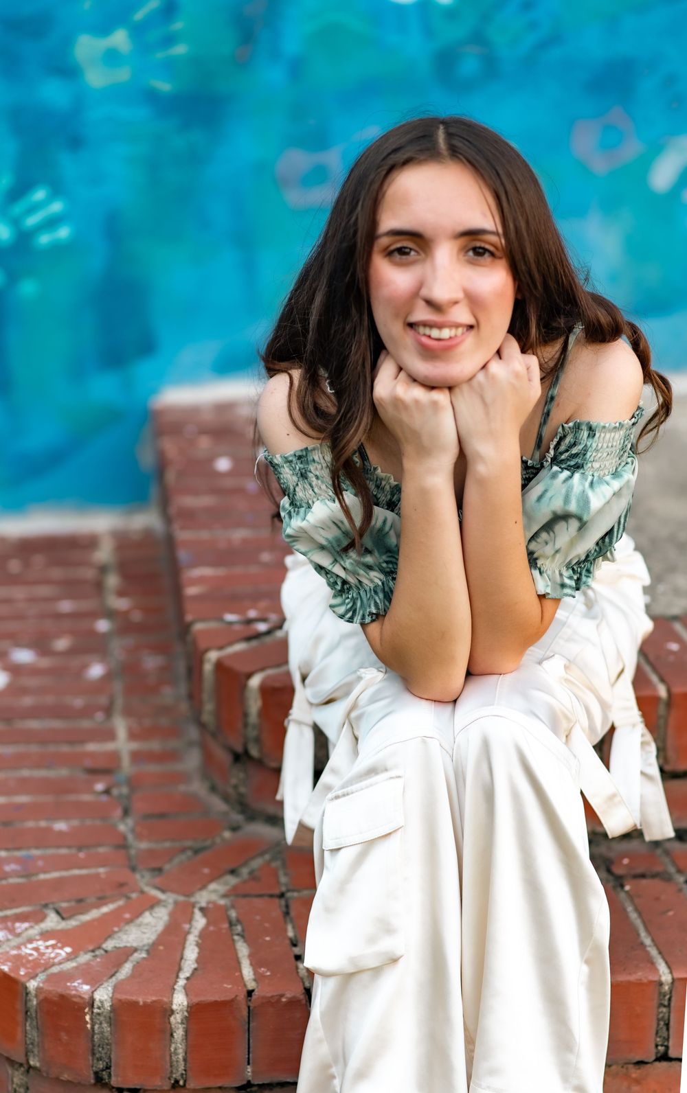 Portrait of graduate in white and green outfit sitting on brick steps with blue backdrop.