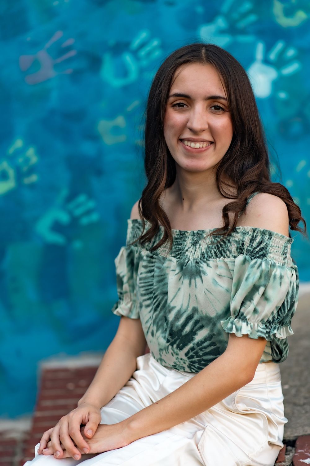 Graduate poses against blue handprint wall wearing off-shoulder green and white outfit.