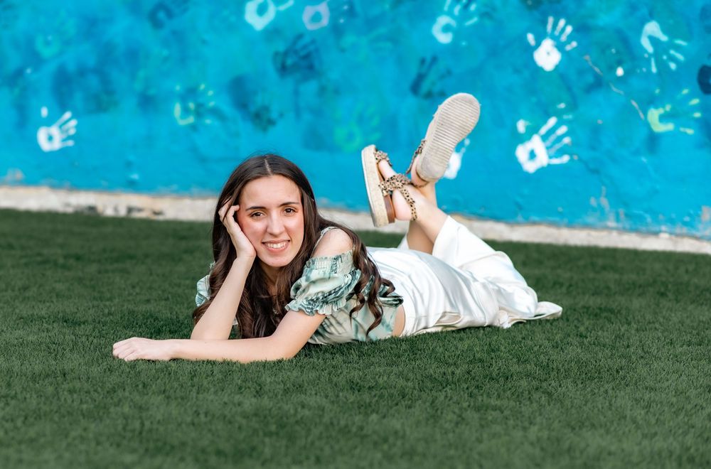 Graduate lying on grass with blue handprint wall background in casual pose.