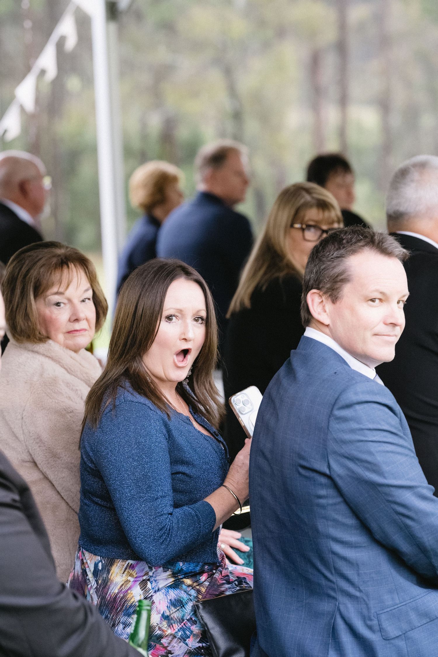 Wedding guests seated at an outdoor venue react with animated expressions during the celebration.