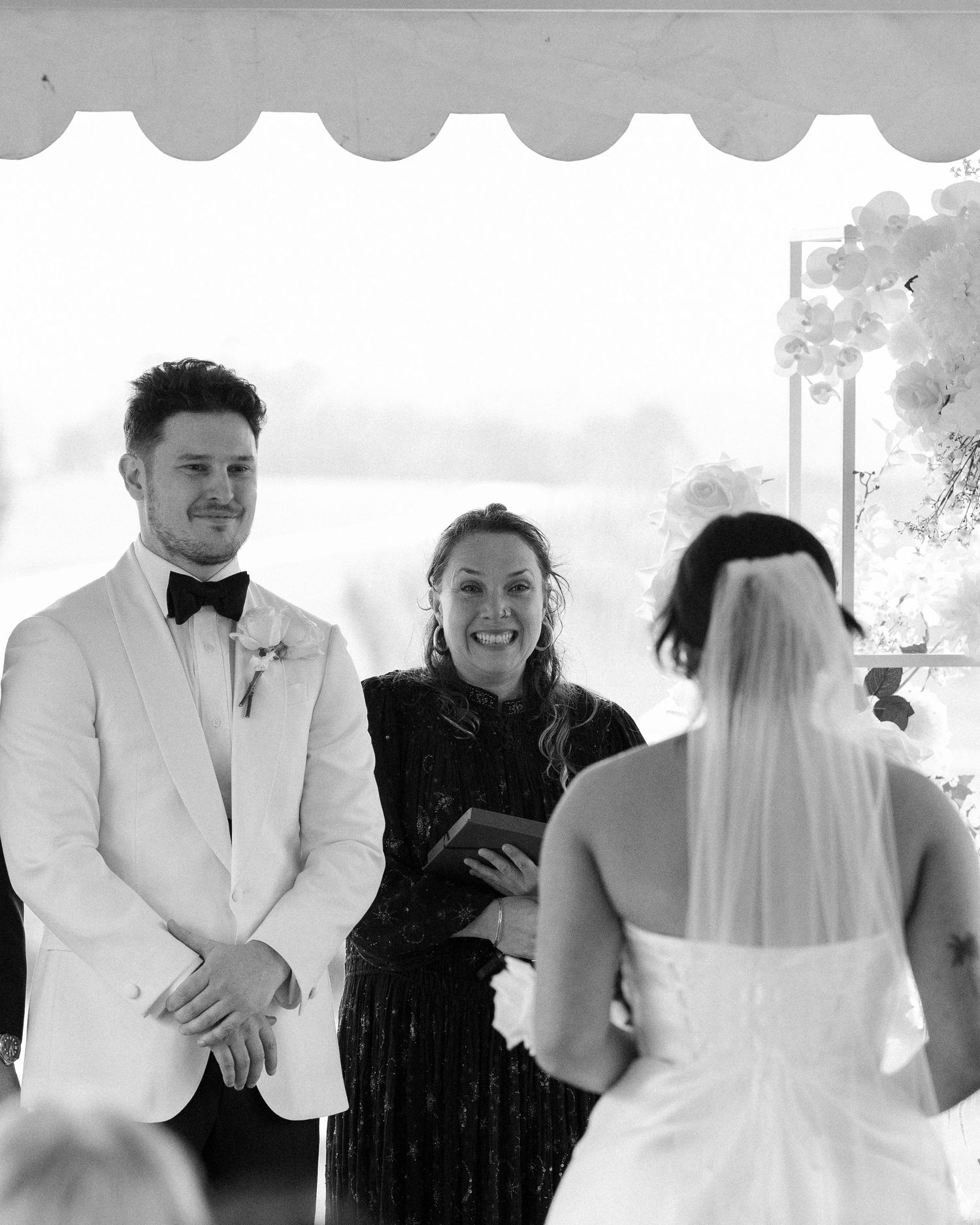 A wedding ceremony taking place outdoors under a canopy with an officiant and couple exchanging vows.