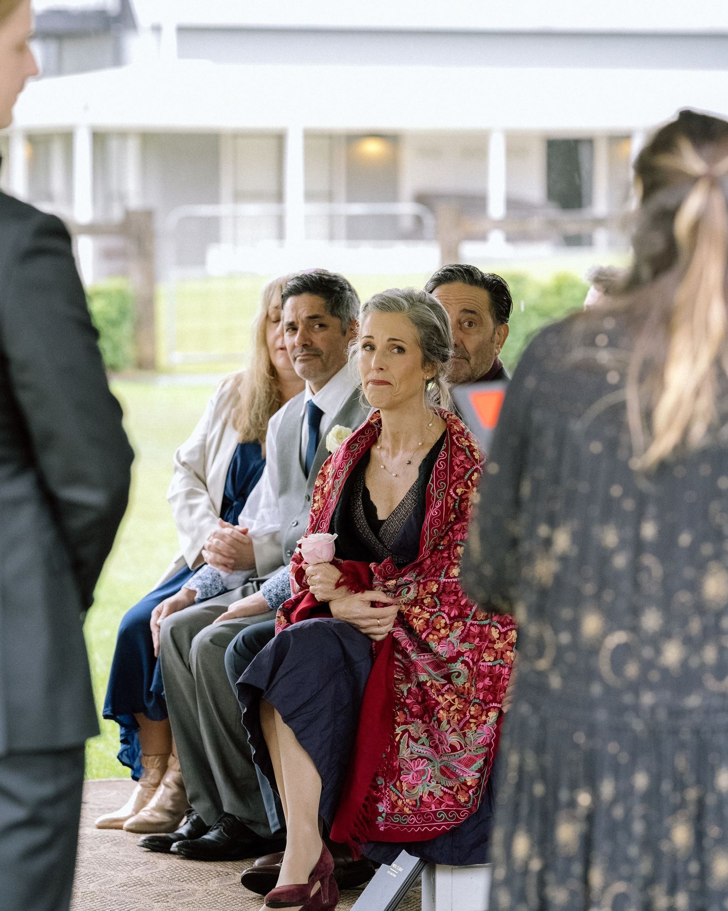 Wedding guests seated on a porch watching an outdoor ceremony with focused expressions.