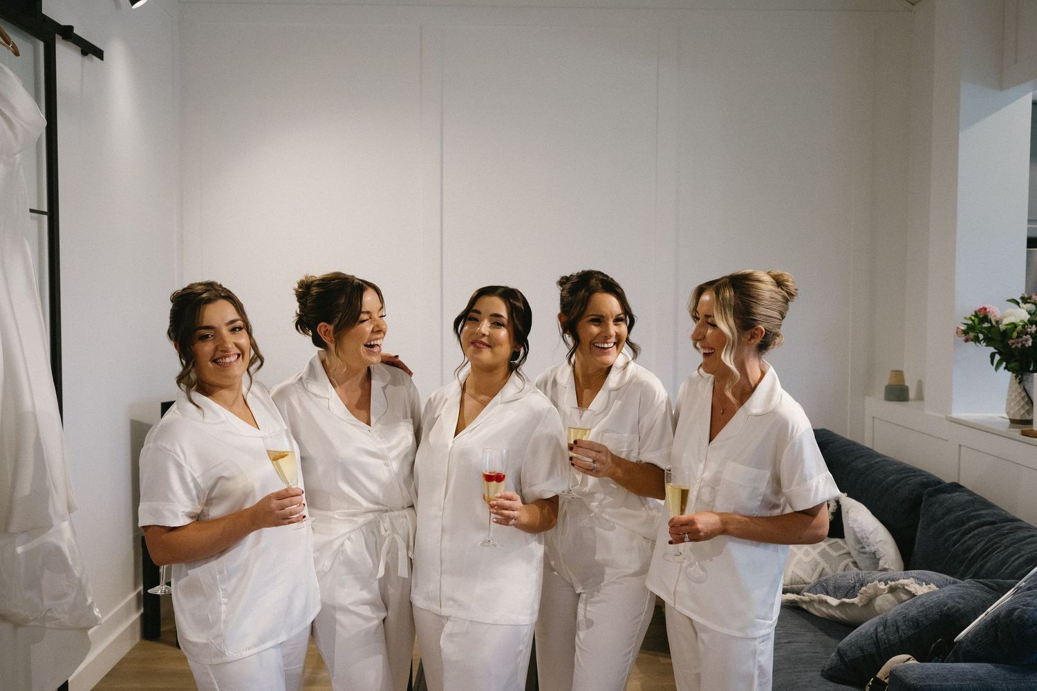 A group wearing matching white robes gather together laughing and holding drinks during pre-wedding preparations.