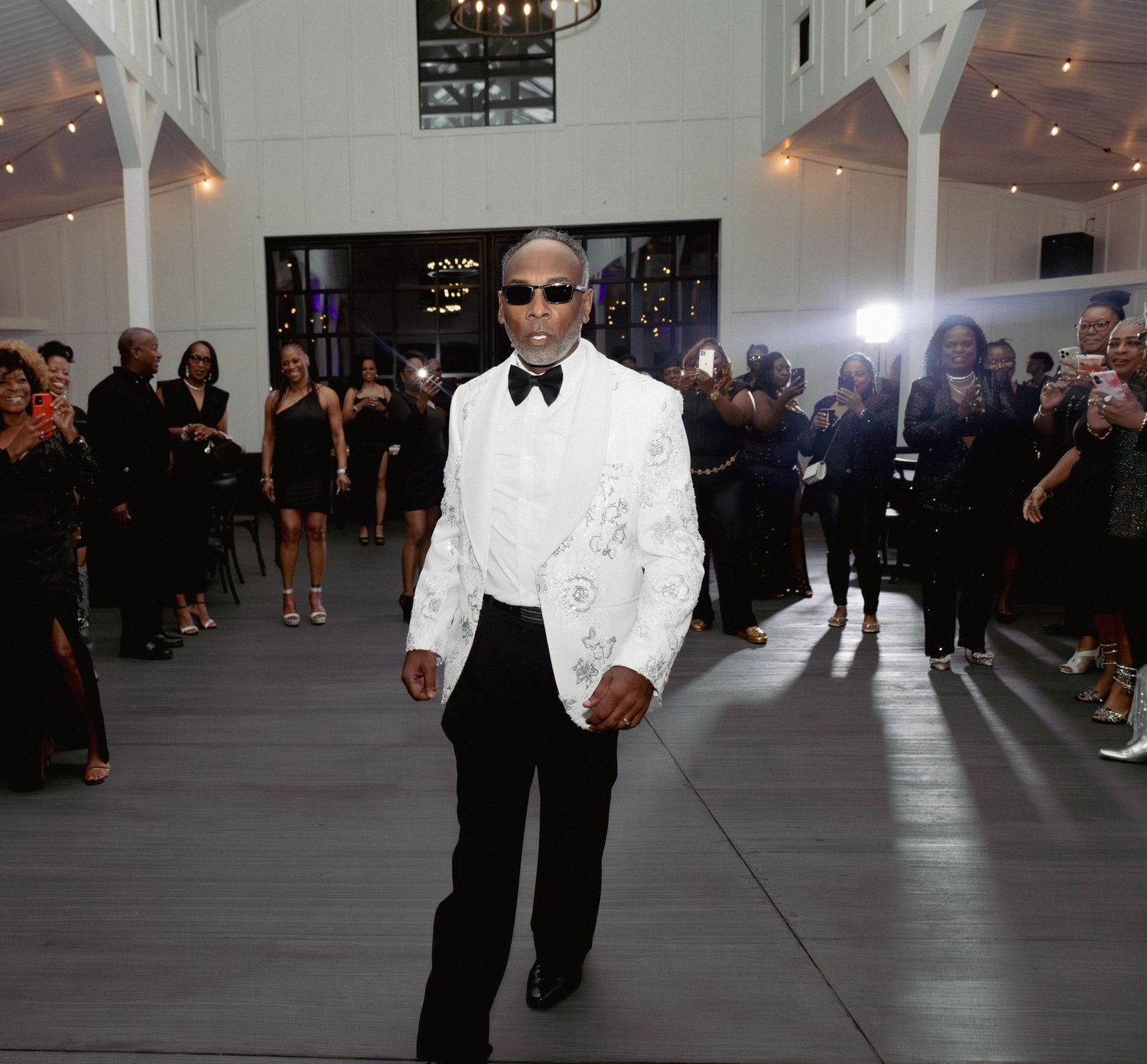Stylish man in white floral blazer and sunglasses walks confidently through elegant white event venue.