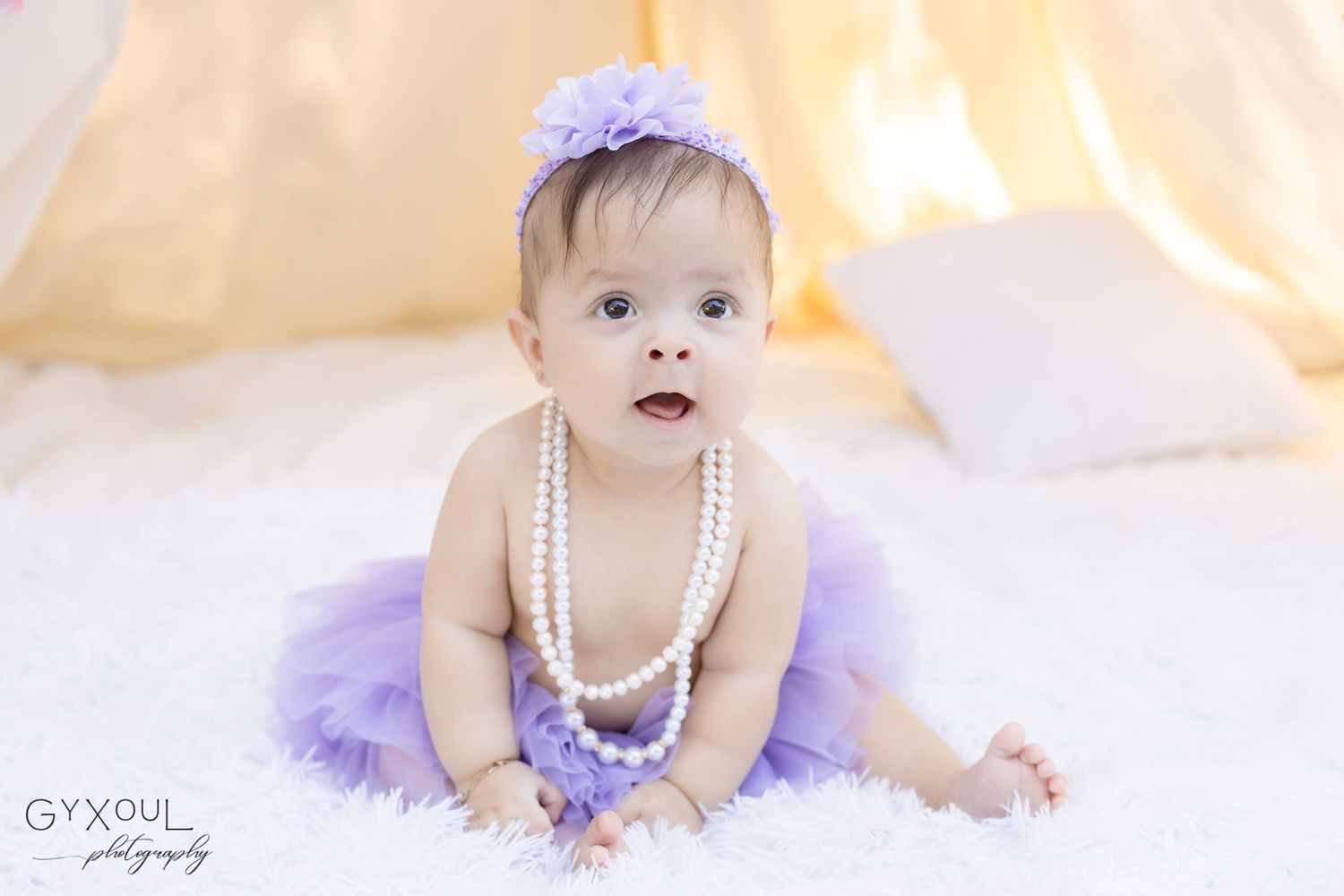 Baby in purple tutu and pearls sitting on white blanket with matching hair bow.