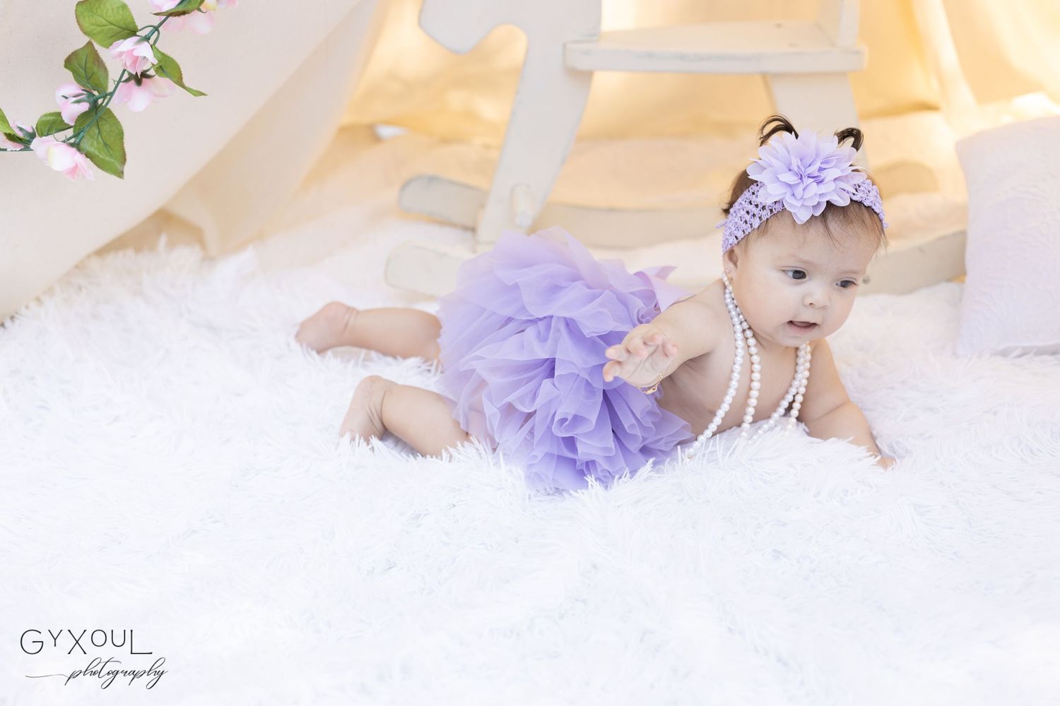Baby wearing purple tulle skirt crawling on white blanket with floral decorations.