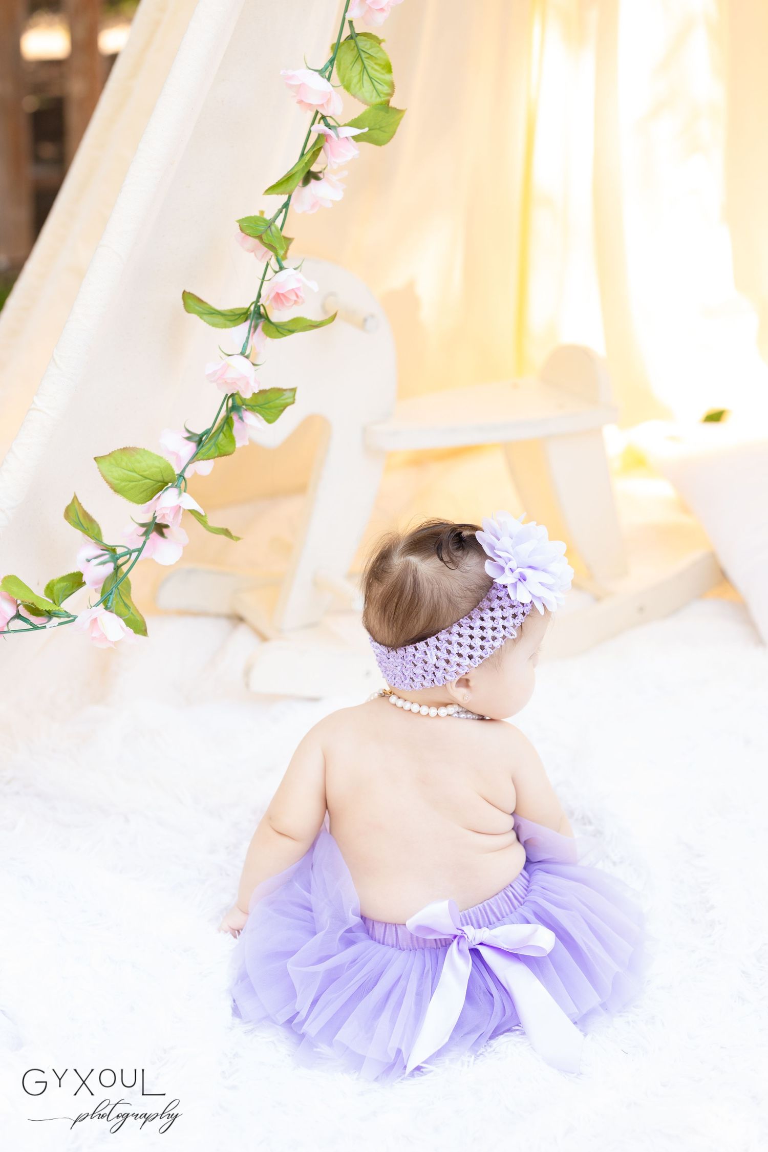 Back view of baby in purple tutu sitting under draped fabric with floral vines.