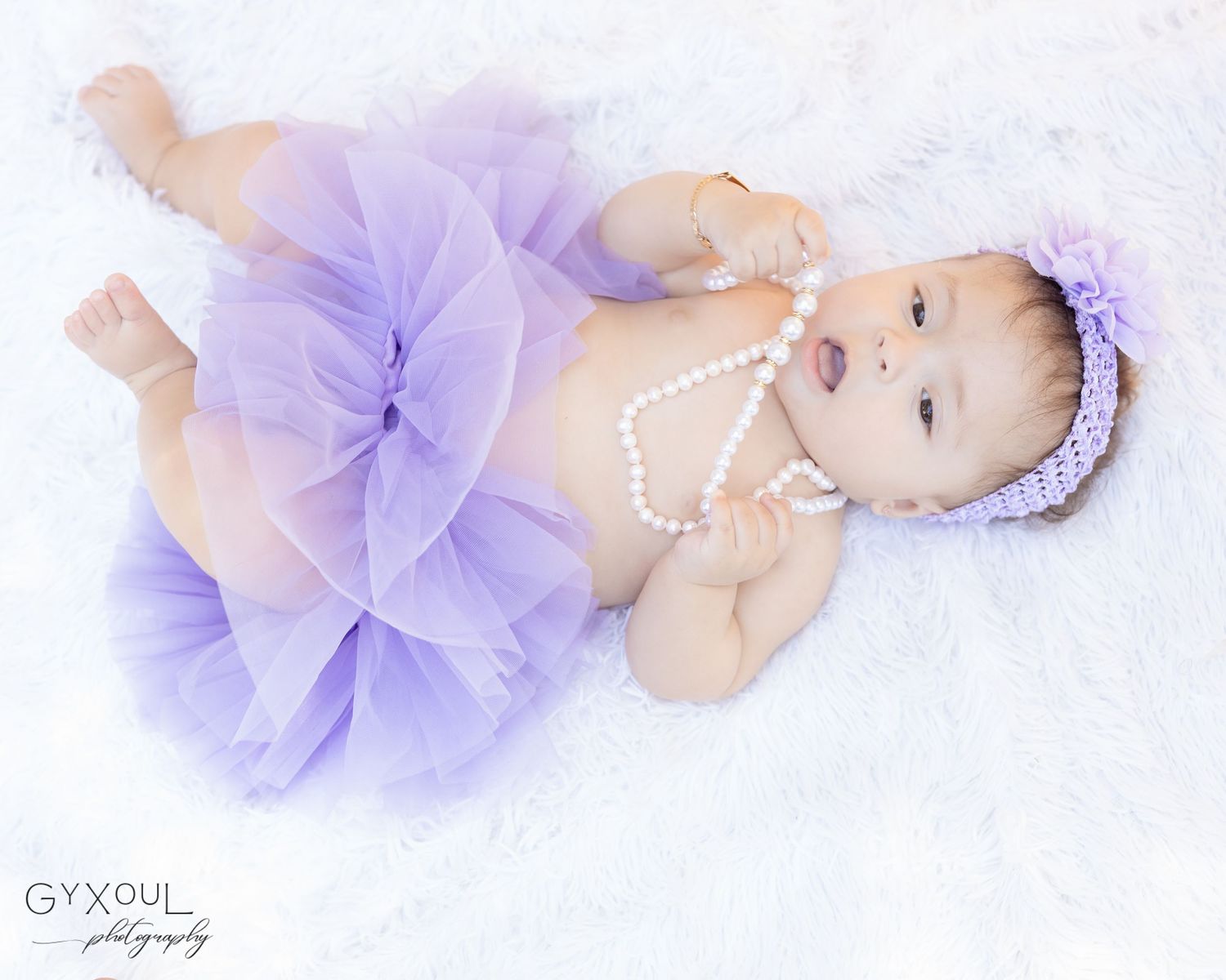 Baby in lavender tulle dress and pearls lying on white fluffy backdrop.