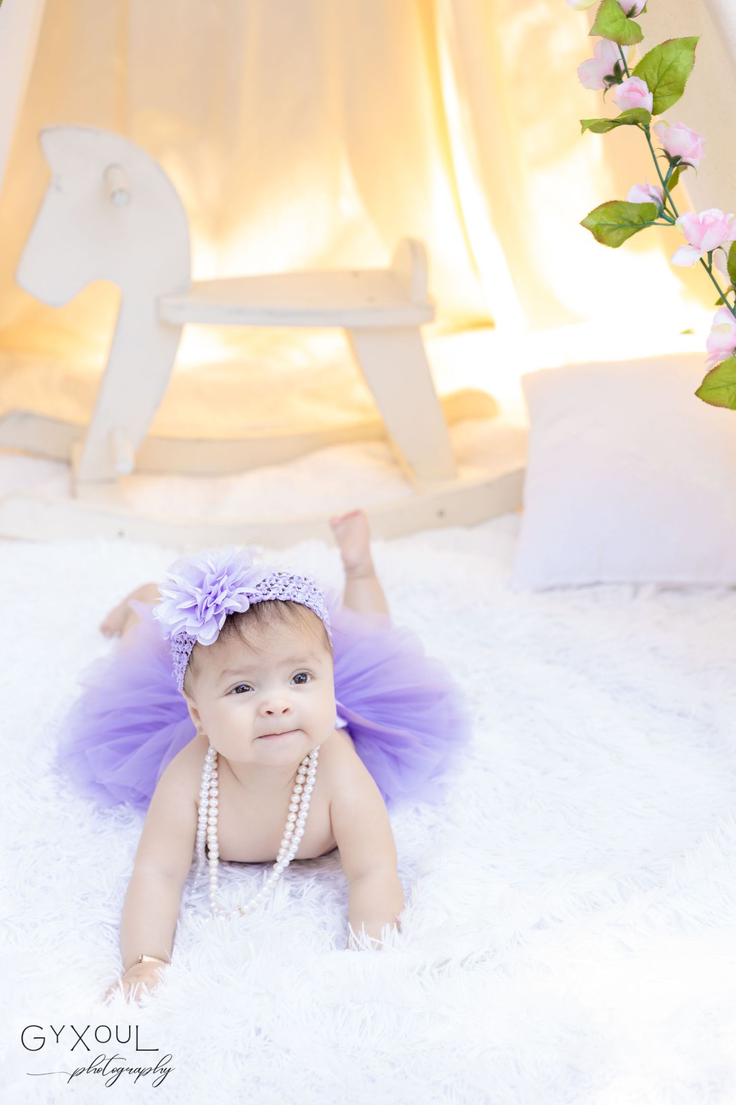 Baby in purple tutu dress near white rocking horse with floral accents.