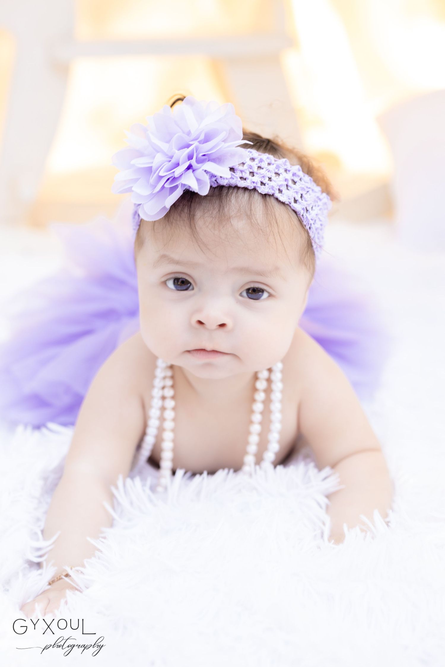 Baby wearing purple headband and tulle dress lying on white fur backdrop.
