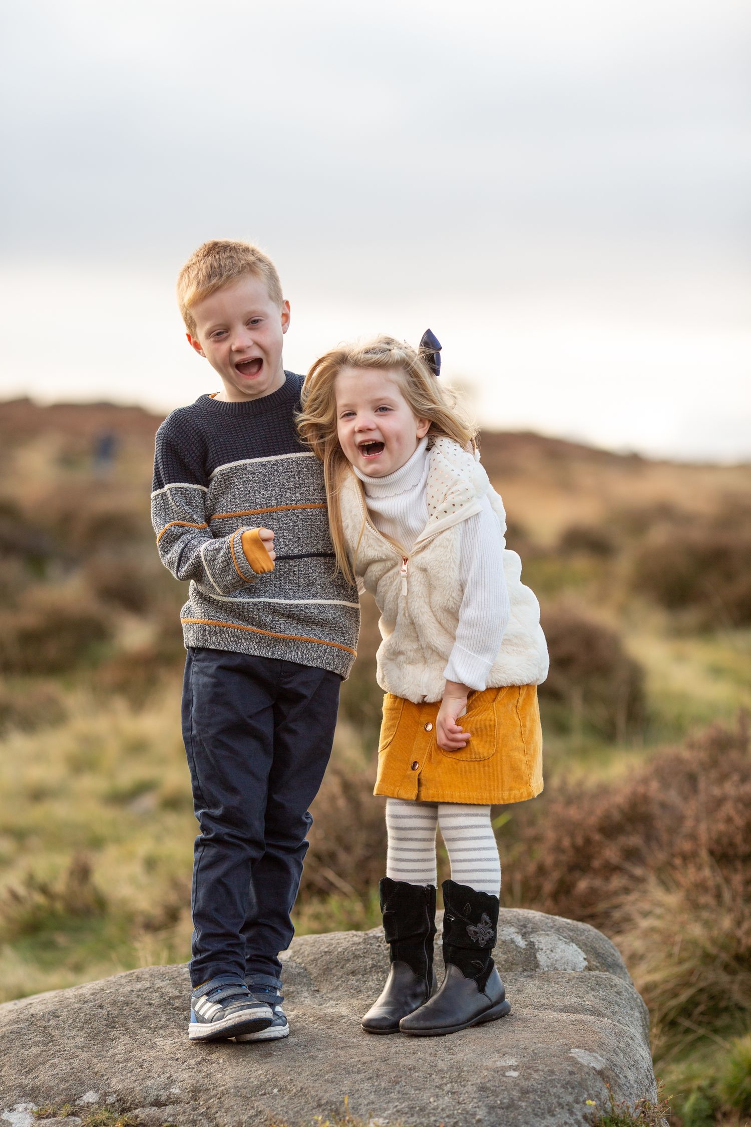 Two children laughing together while standing on rock in countryside setting.