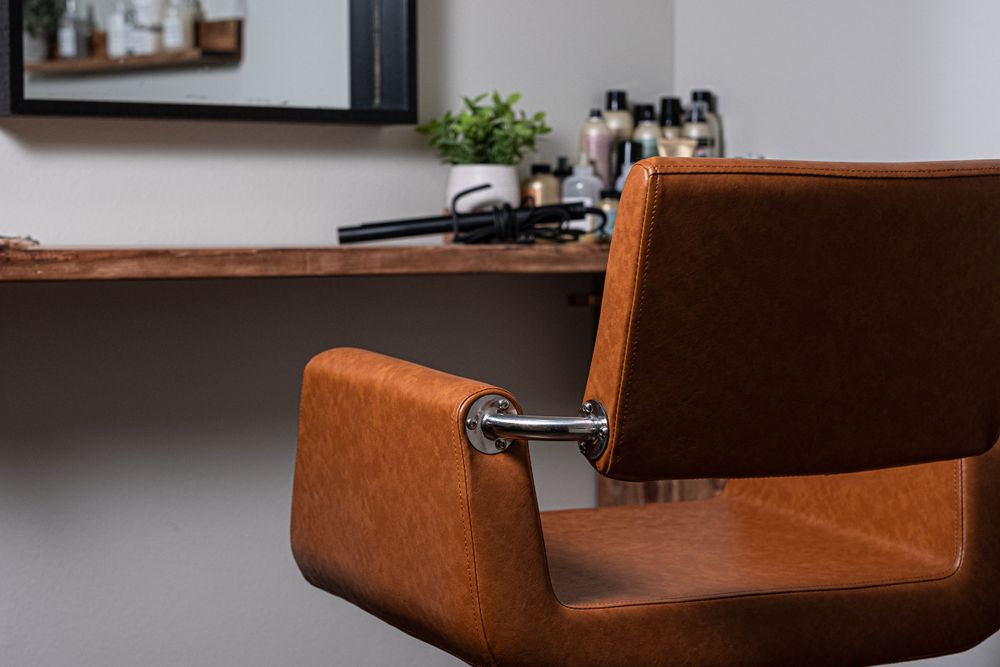 Vintage brown leather barber chair shown against gray wall with wooden floating shelf in background.