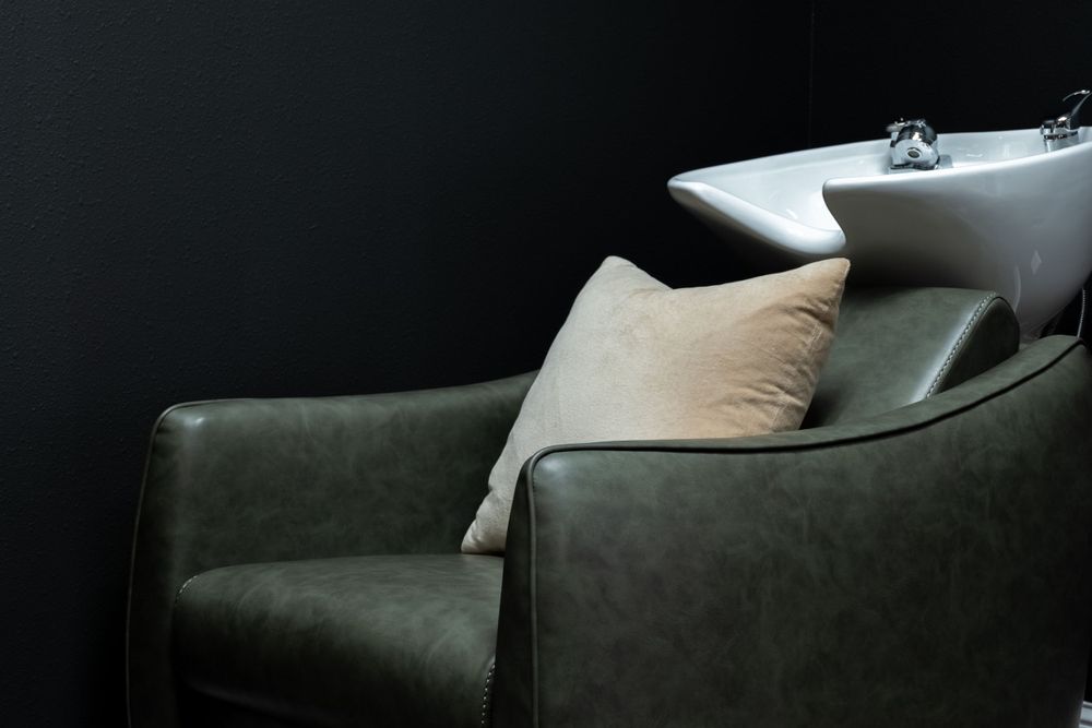 Hair salon washing station with dark green leather chair and white ceramic sink basin against black background.