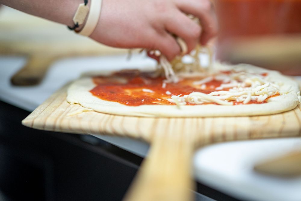 Hand sprinkling shredded cheese onto raw pizza dough covered with tomato sauce on wooden pizza peel.