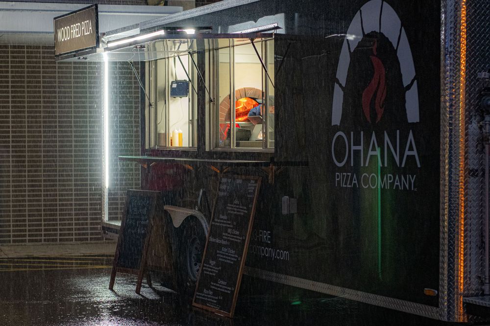 Rainy night view of Ohana Poke Company storefront with illuminated window and chalkboard menu sign outside.