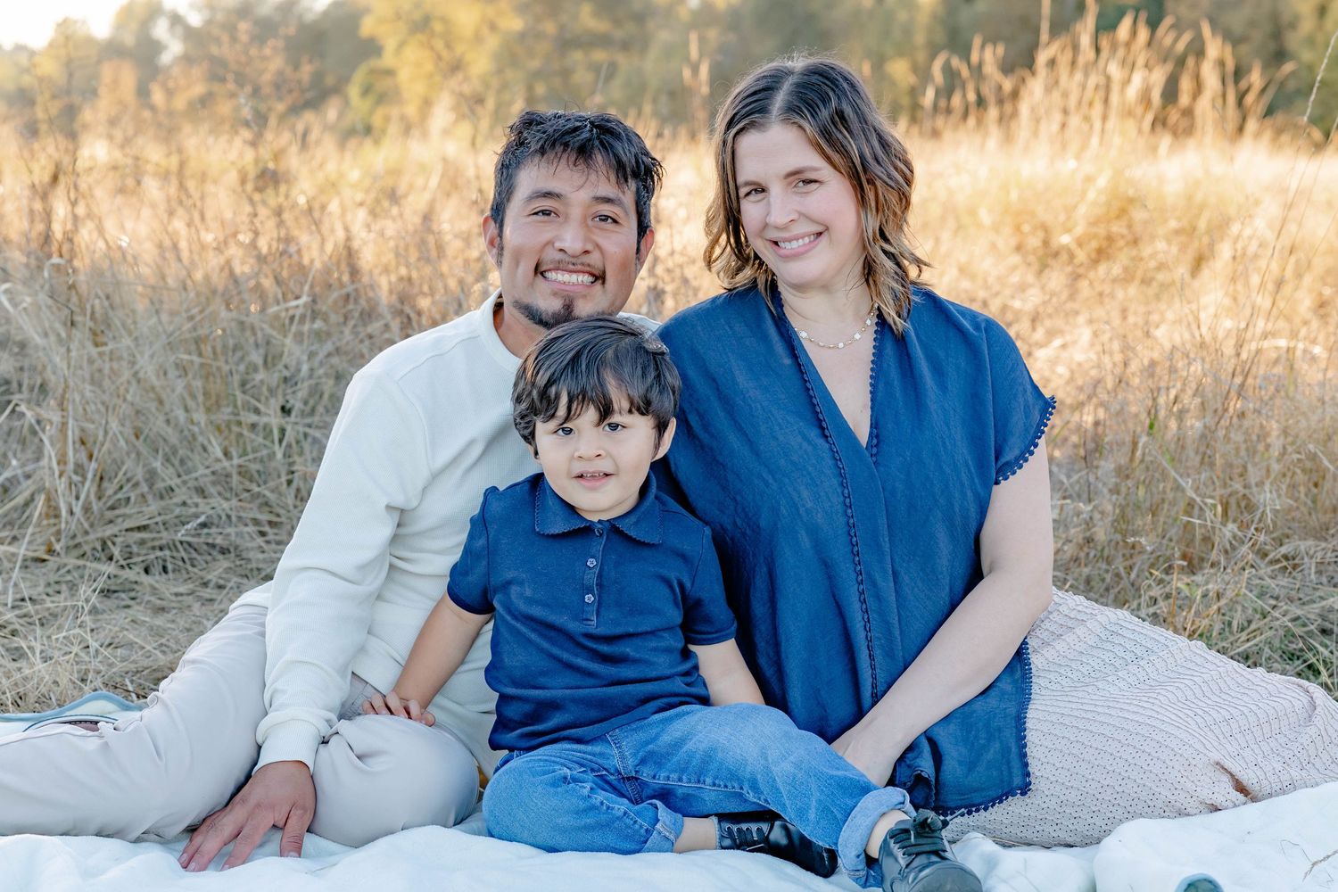 Smiling family of three sits together on a blanket in a golden grass field at sunset.
