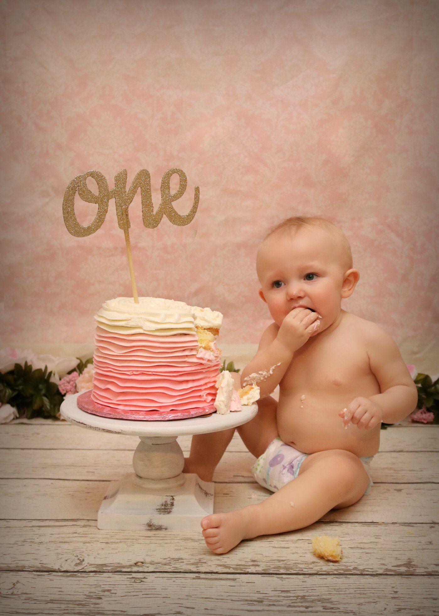Baby in diaper enjoying pink ombre cake with gold 'one' topper on wooden floor.