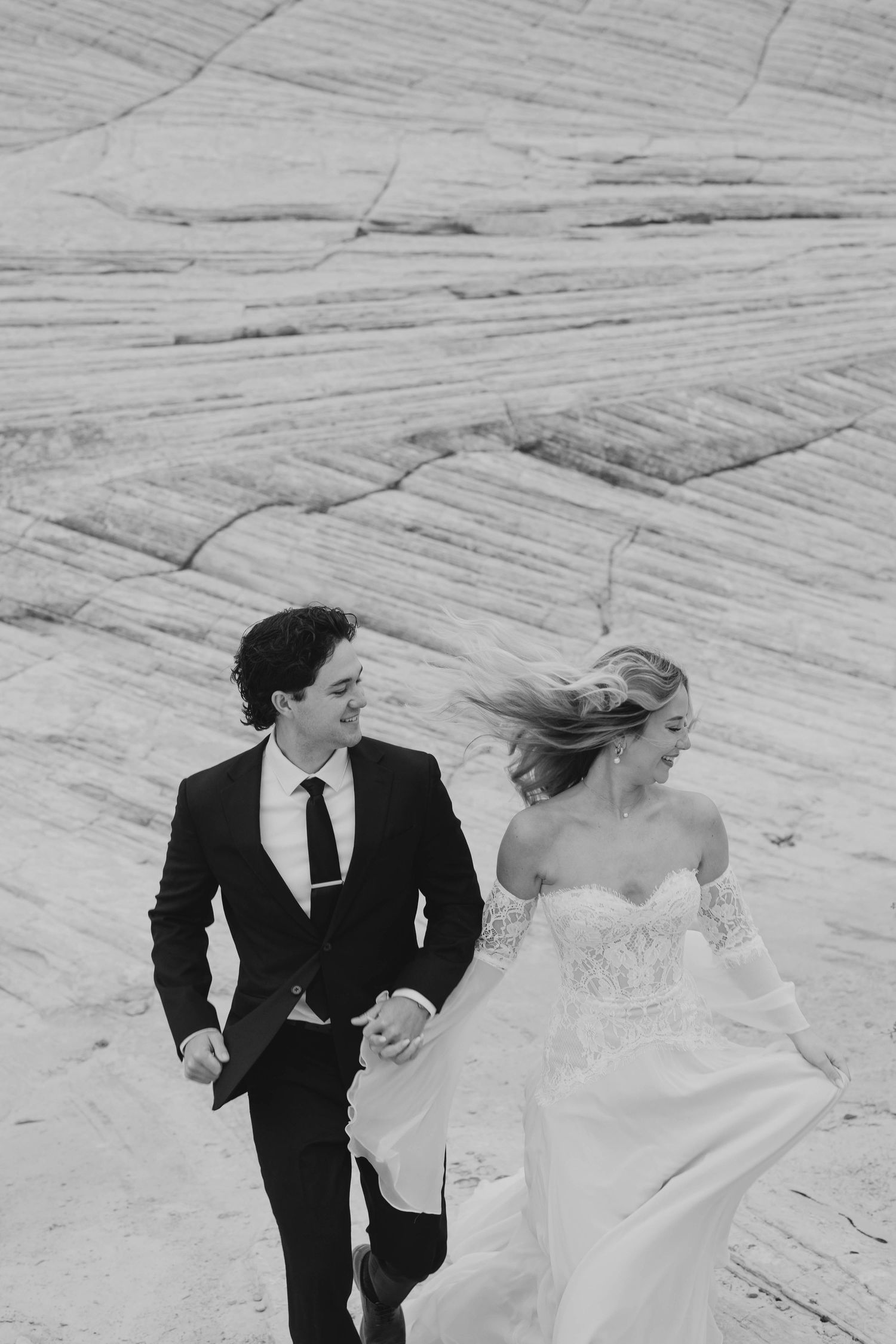 A bride and groom run hand in hand across dramatic white rock formations in a black and white wedding photo.