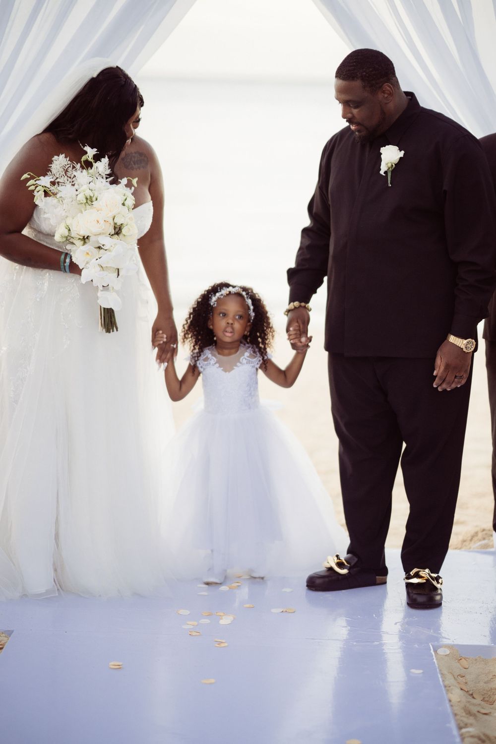 Family in formal attire during beach wedding ceremony under white fabric canopy.