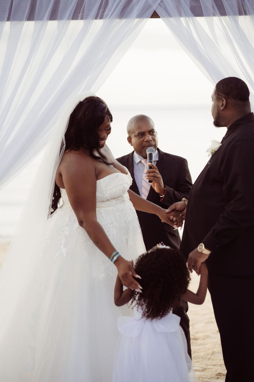 A beach wedding ceremony with a couple exchanging vows under white drapes as a small child watches.