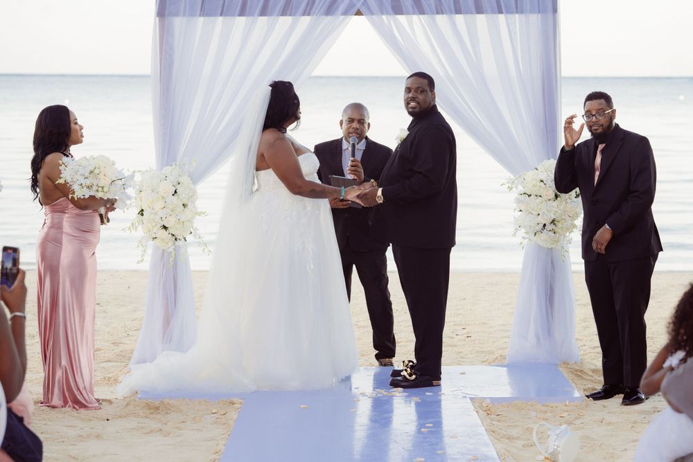 A wedding ceremony on a blue aisle runner with white drapes and floral arrangements on the beach.