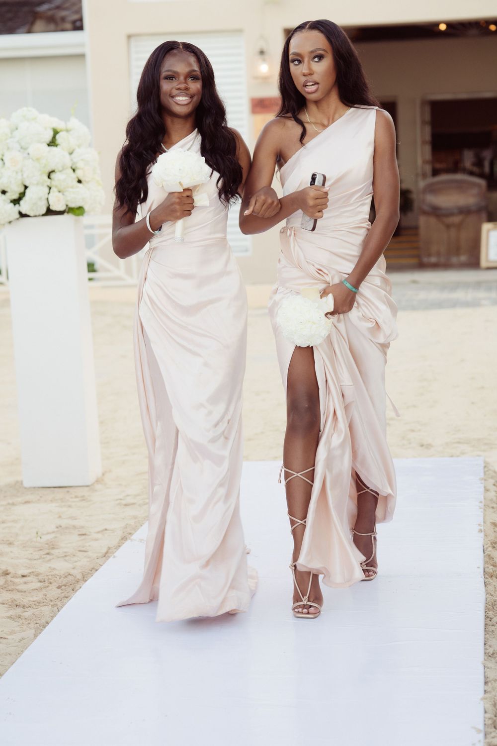 Two bridesmaids in elegant blush gowns walking down a white aisle runner at an outdoor wedding venue.
