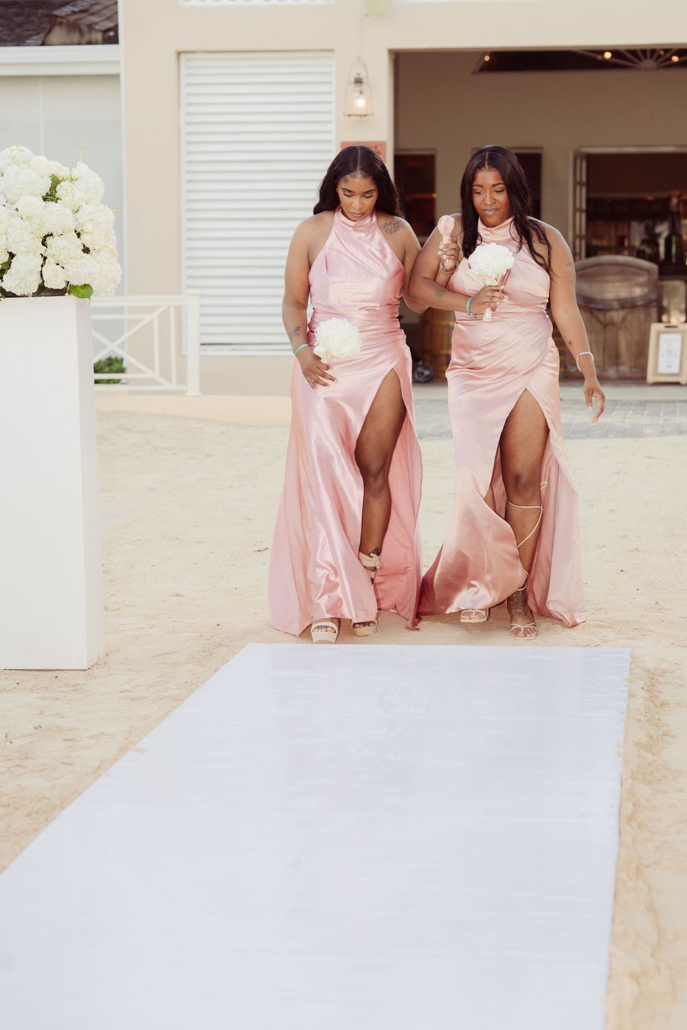 Bridesmaids in pink satin dresses with high slits walk down a white aisle runner.