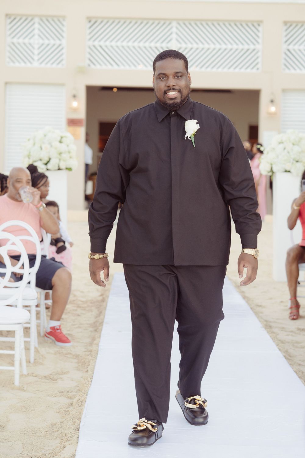 A groom in a black suit walks down a white aisle runner during an outdoor beach ceremony.