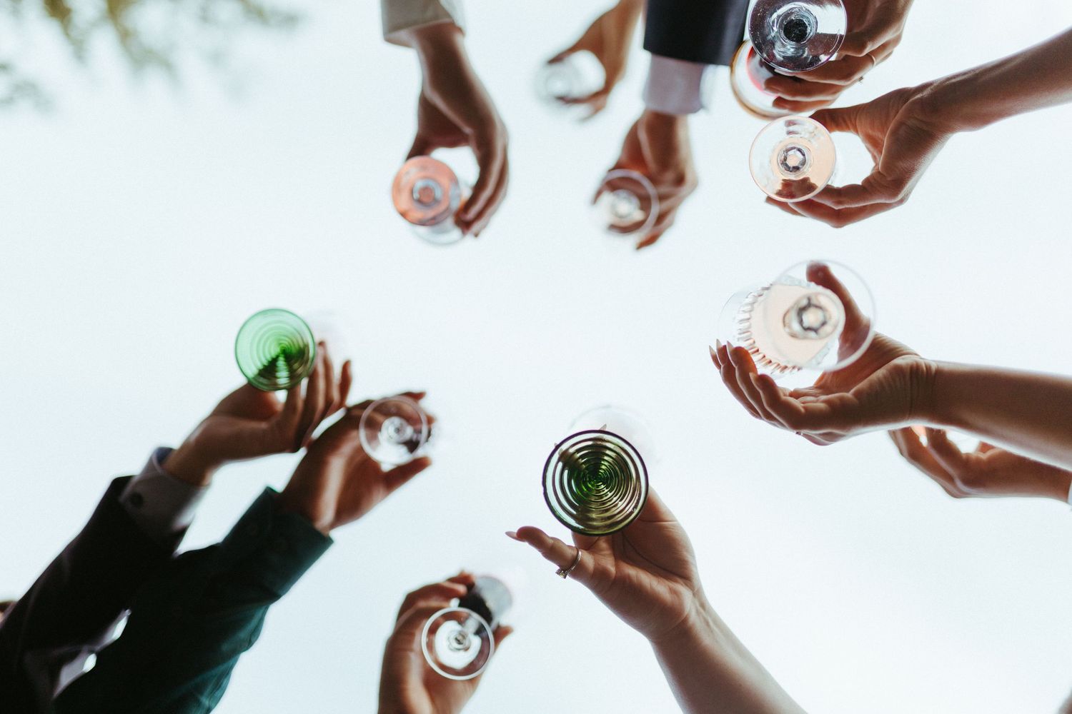 Group of people raising cocktail glasses in a celebratory toast viewed from below against a bright sky.
