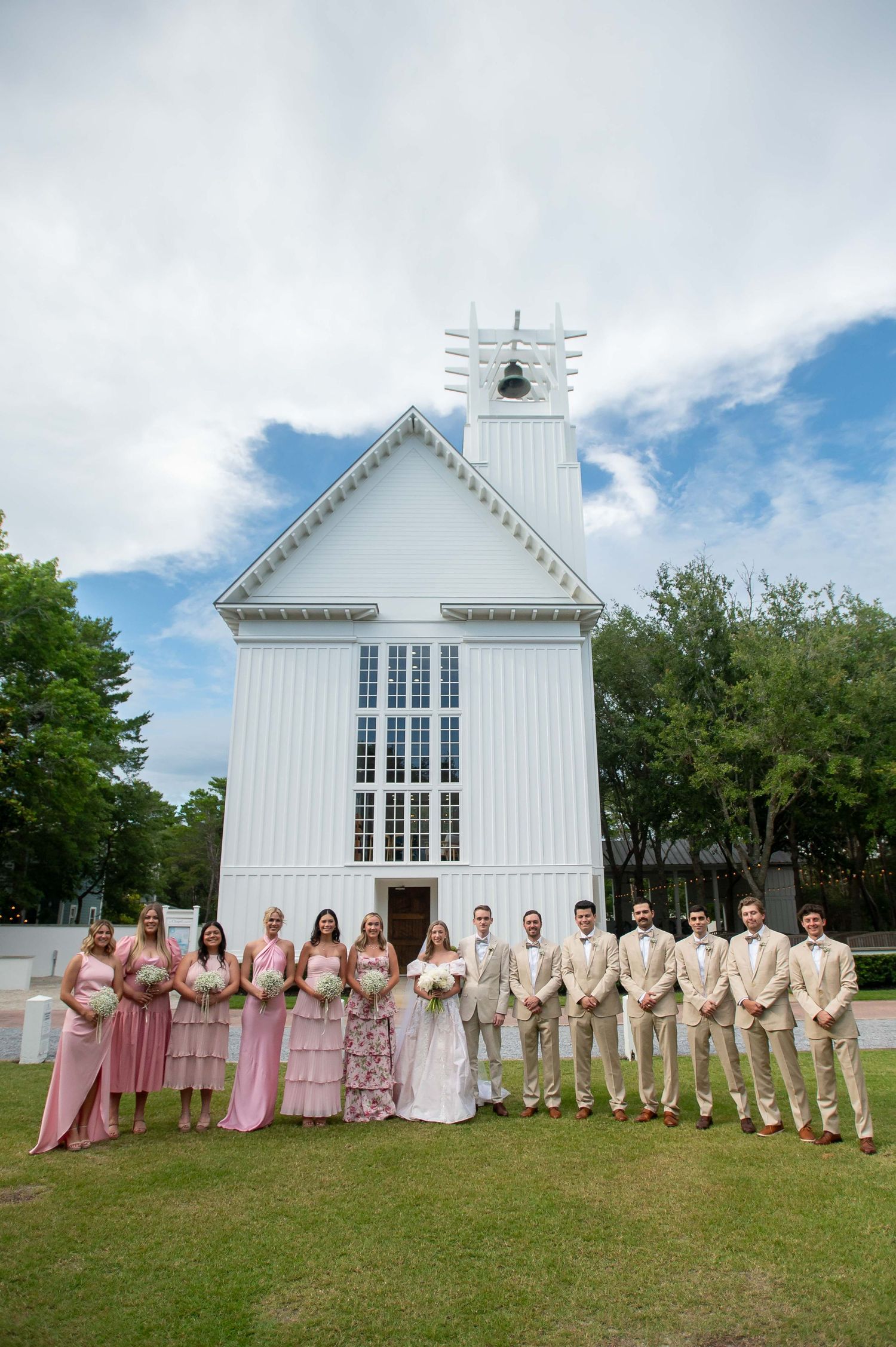 Wedding party in pink and beige attire poses in front of a charming white chapel with tall windows under blue skies.