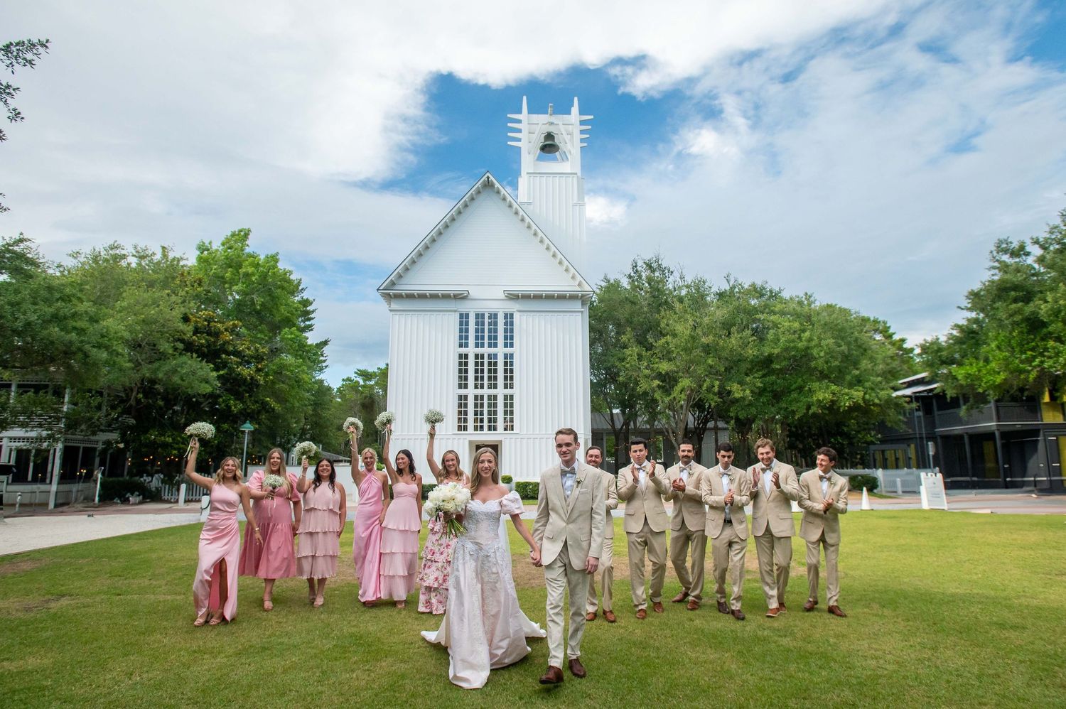 Wedding party walks away from small white chapel on sunny day with bridesmaids in pink and groomsmen in tan suits.