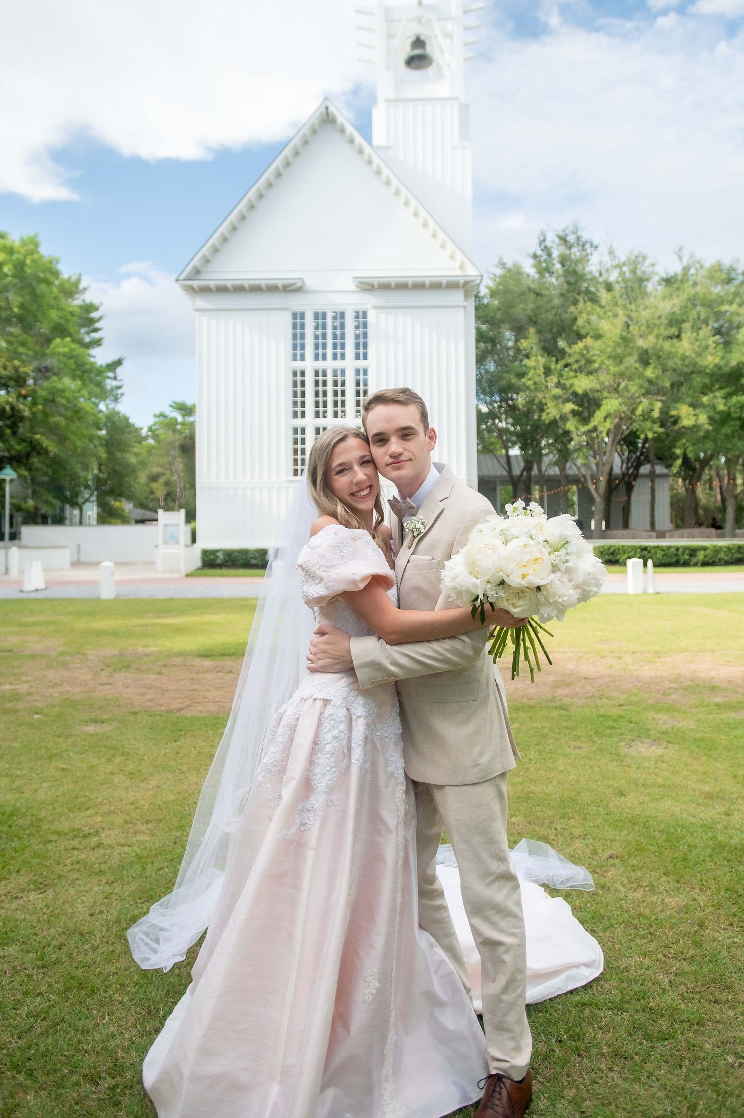 A newlywed couple embraces in front of a beautiful white chapel with tall steeple on a sunny summer day.