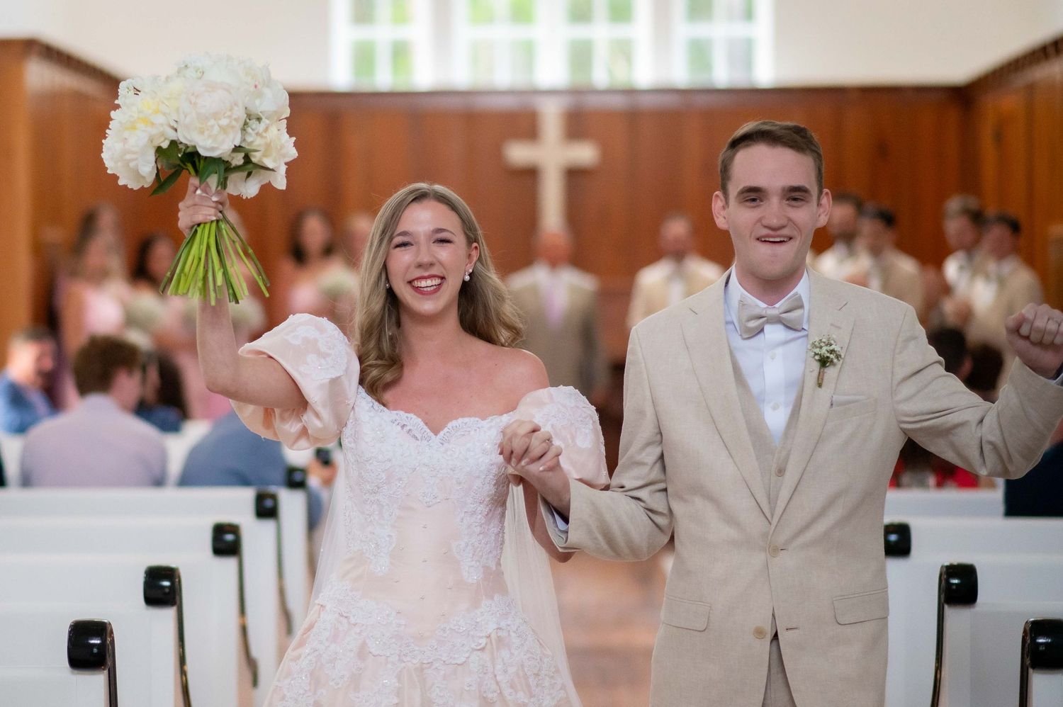 Joyful couple celebrates at the altar of a traditional church ceremony with white bouquet and tan suit.