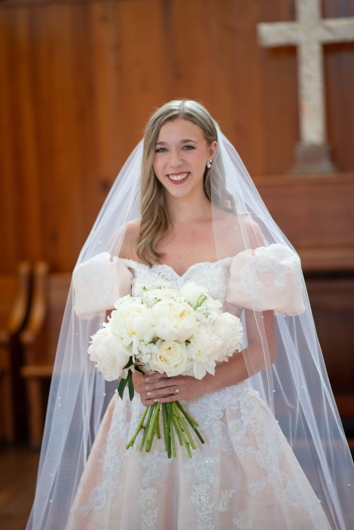 A bride in a white lace wedding gown and veil holds a bouquet of white peonies in front of a church cross.