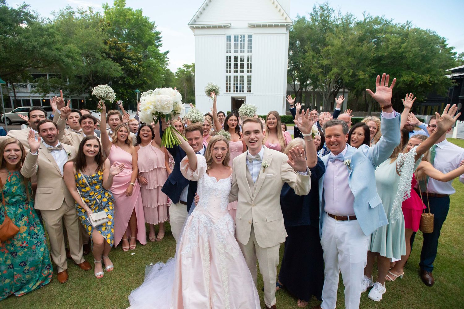 Wedding guests in pink, beige, and pastel attire gather outdoors for a celebratory group photo near a white building.