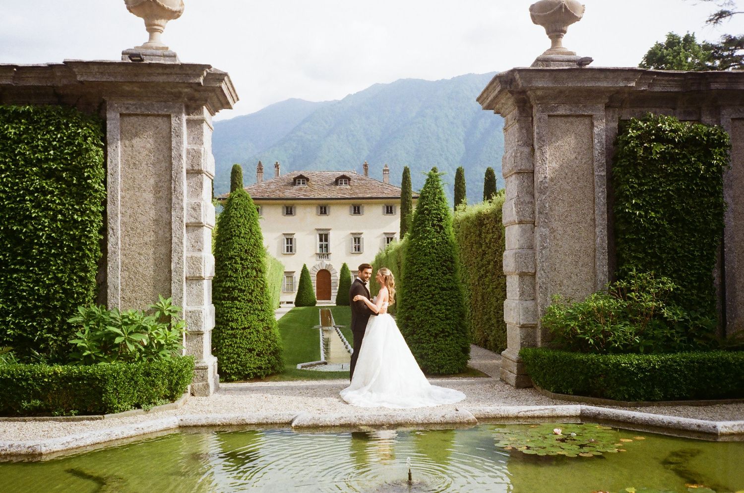 Two people stand beside a reflecting pool framed by stone pillars and manicured hedges at an elegant Italian villa estate.