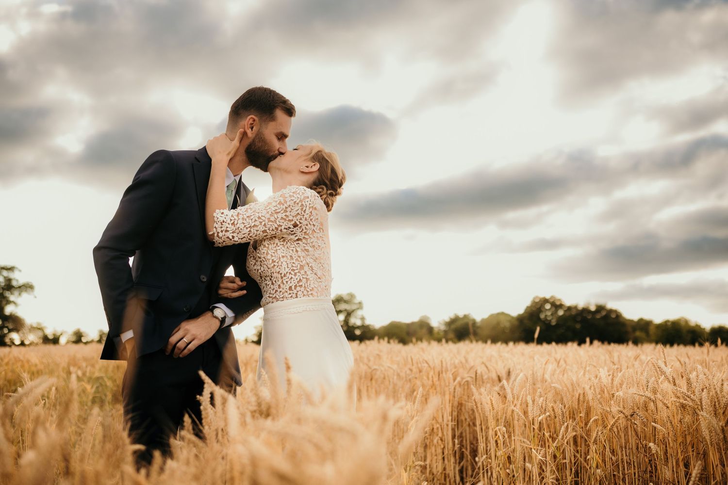 Romantic couple embraces in wheat field under dramatic cloudy sky.