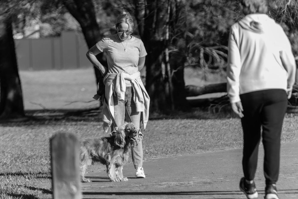 A person in casual wear stands on a path with a small dog while another person walks nearby in a park setting.