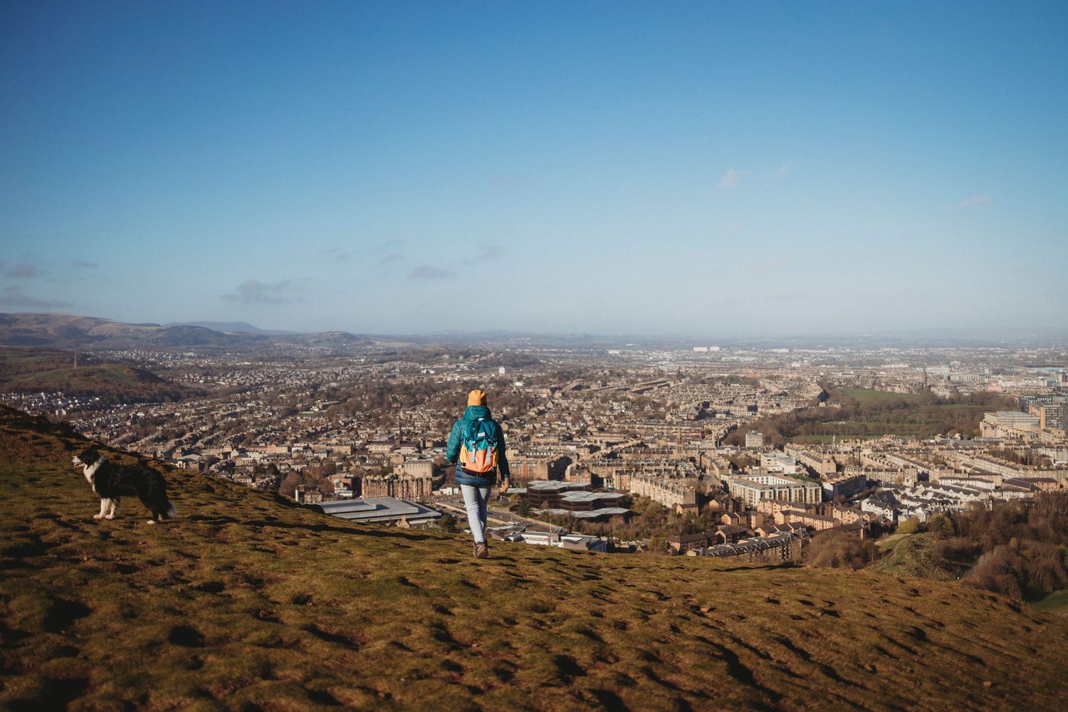 A hiker with a backpack stands on a hilltop overlooking a vast cityscape with mountains in the distance on a clear day.