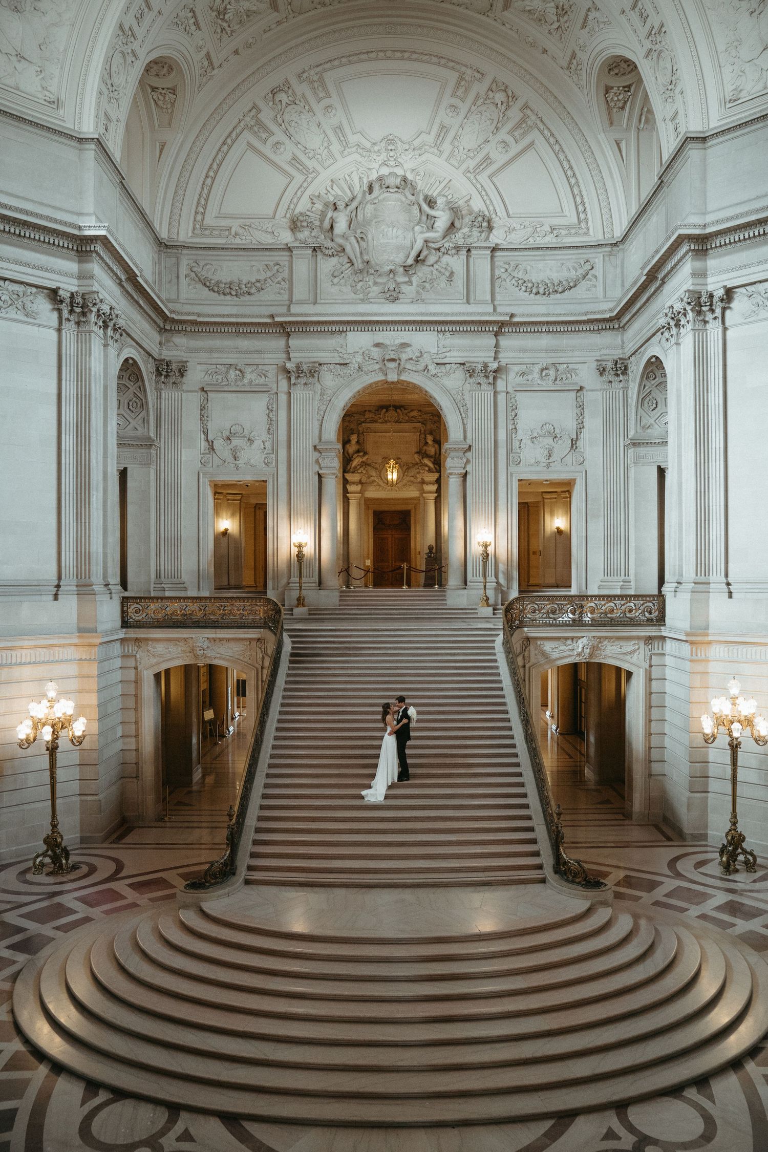 Two figures ascend an ornate marble staircase inside San Francisco City Hall's grand Beaux-Arts style interior.