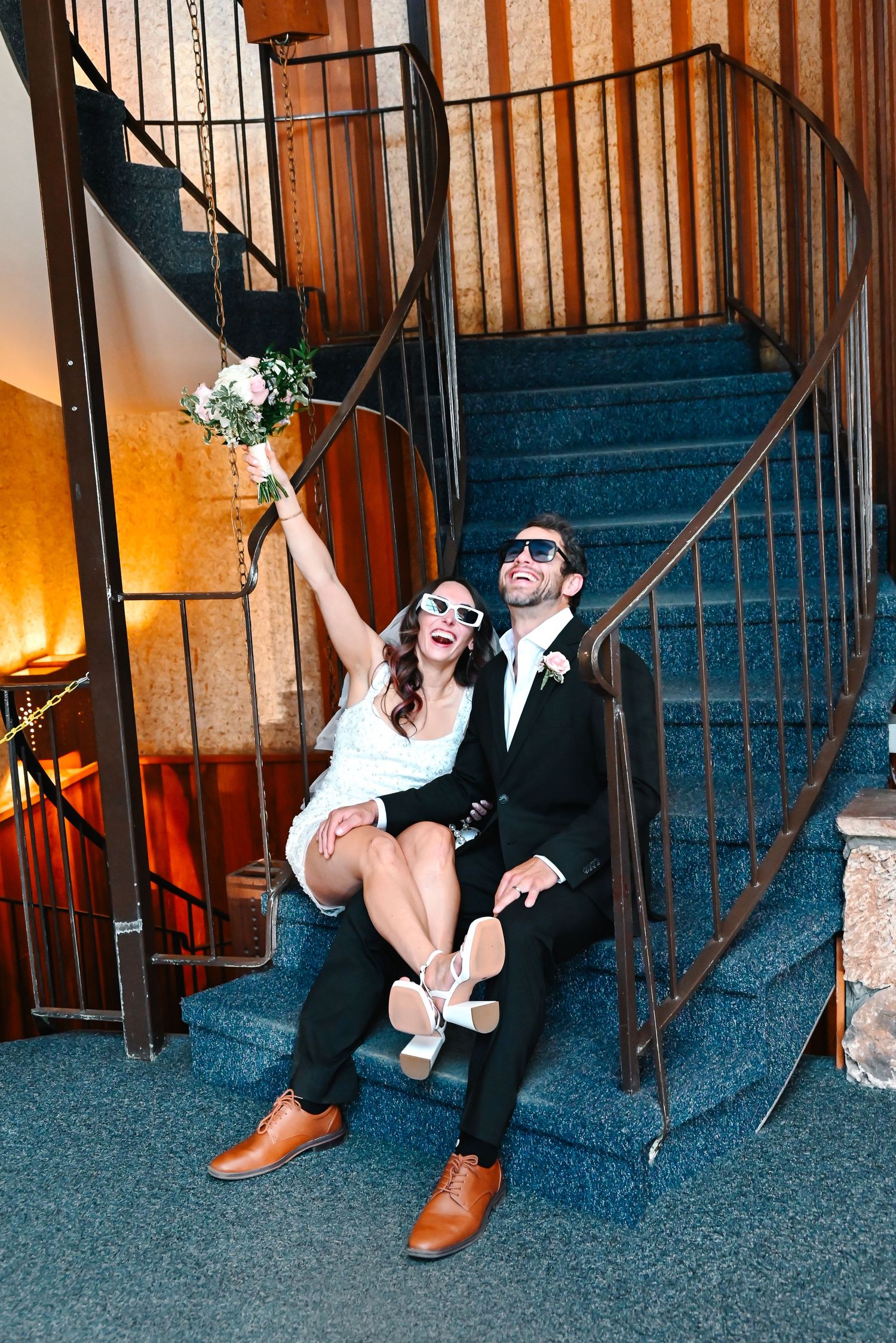 A couple in wedding attire sits on carpeted stairs, with the bride holding up a bouquet while wearing sunglasses.
