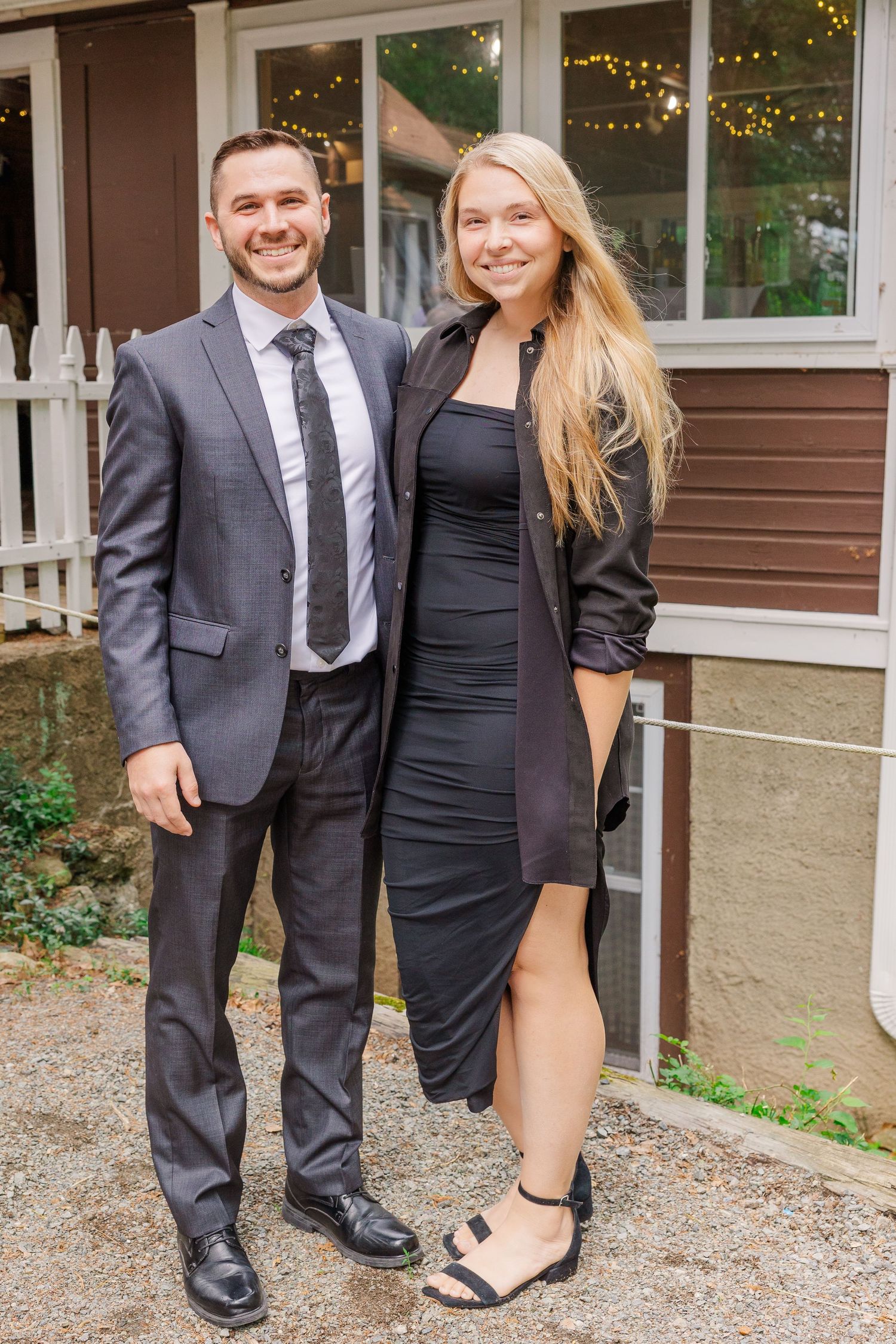 A couple in formal attire poses in front of a brown building with string lights visible in the window.