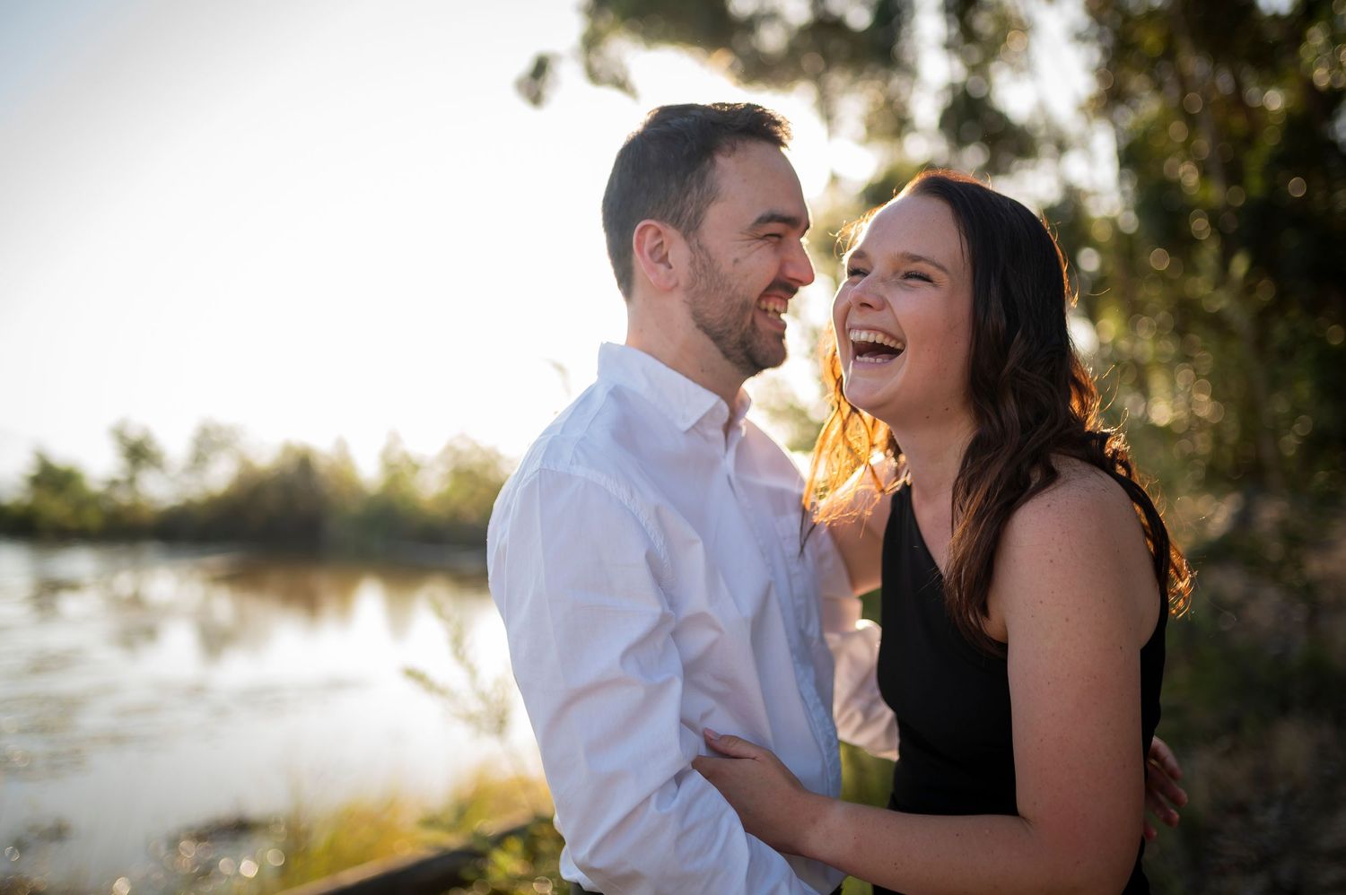 A romantic sunset engagement photo with a couple laughing together by a lake.