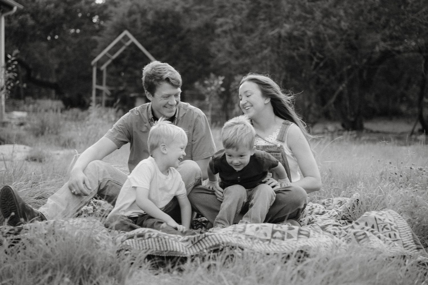 Black and white candid photo of a happy family sitting together on a blanket outdoors in a natural setting.