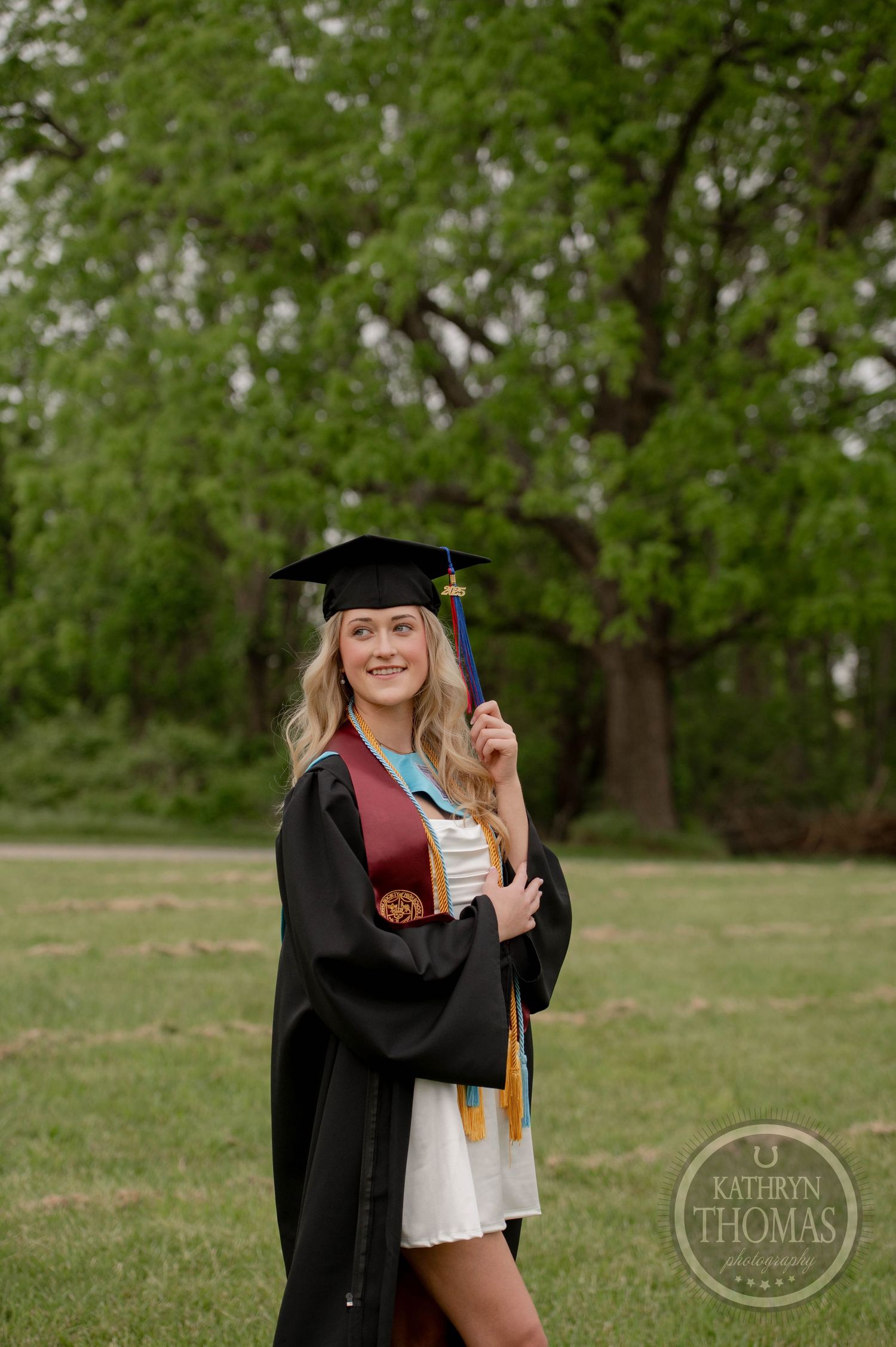 Graduate in black cap and gown poses for photos on green grass field during graduation photoshoot session.