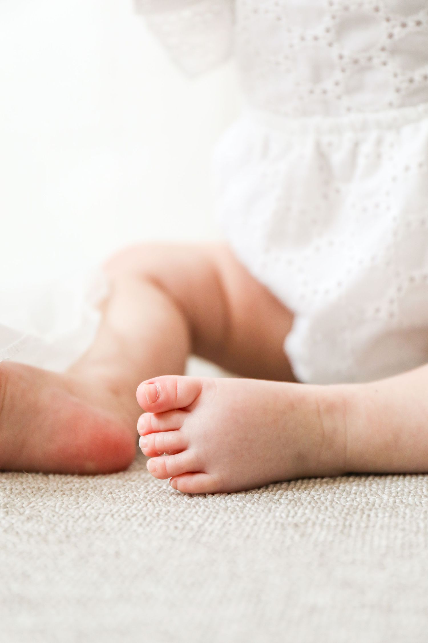 Close up view of a baby's tiny feet peeking out from white eyelet fabric while resting on beige carpet during Birthday shoot in Nottingham