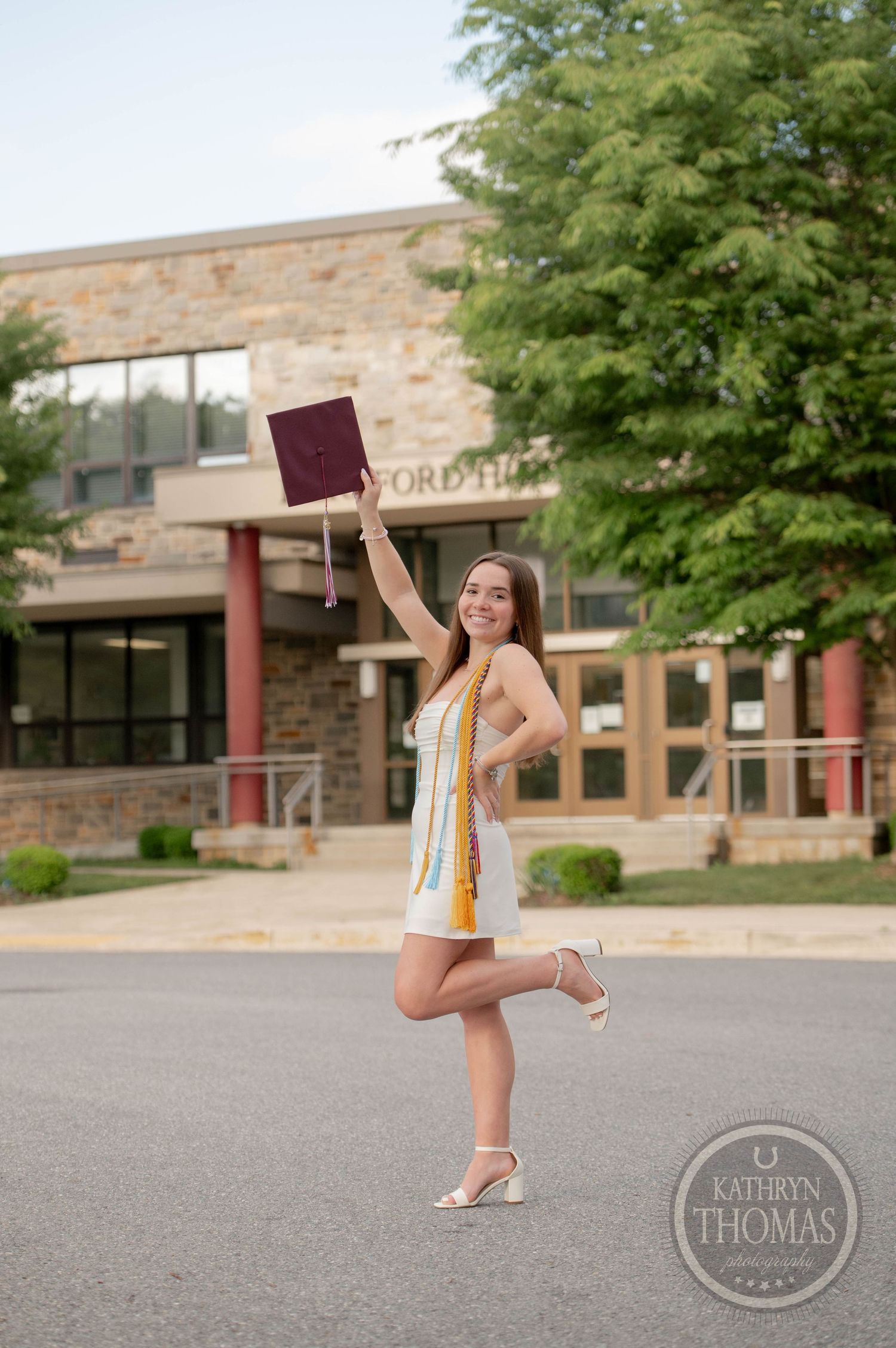 Graduate in a yellow sundress celebrates with diploma in front of brick building, striking a joyful pose.