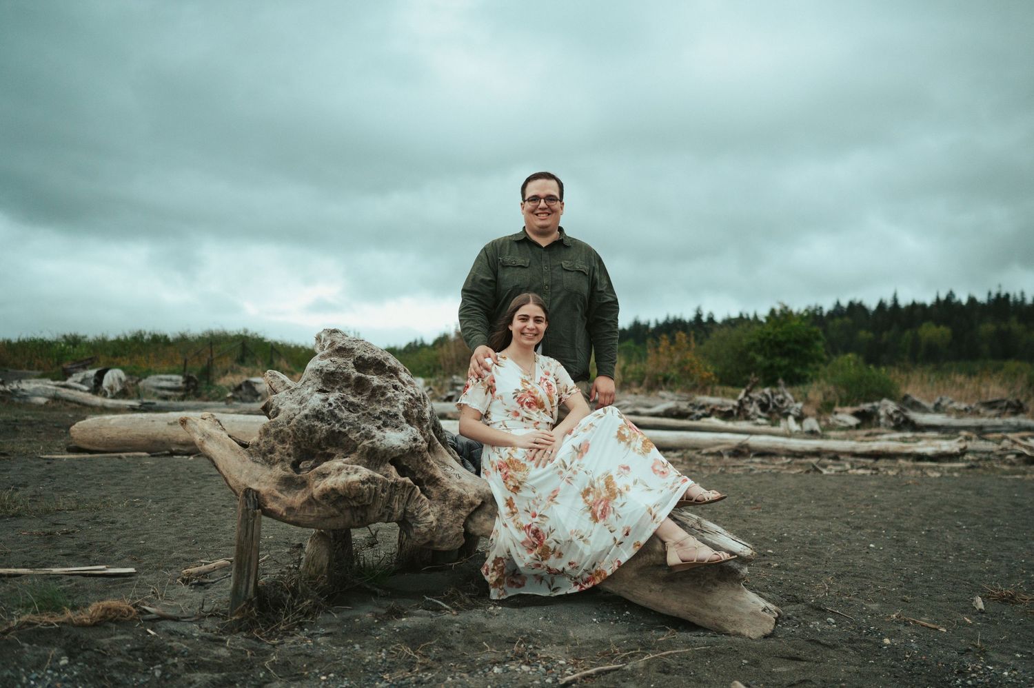 A couple poses on driftwood at a moody beach with dark cloudy skies and forest in the background.