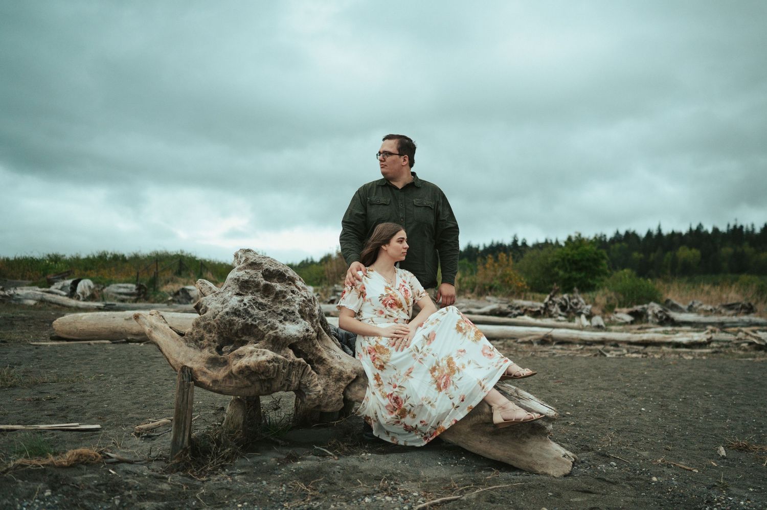 A couple sits together on driftwood at a moody beach with cloudy skies and forest in the background.